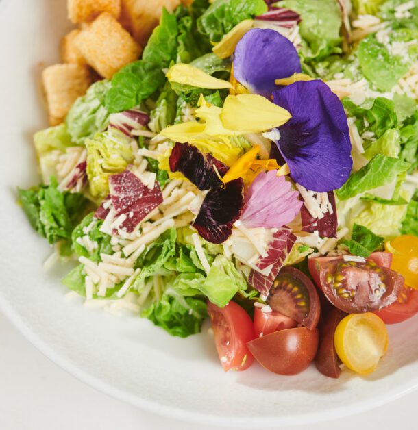 white plate with salad that has tomatoes croutons and a purple flower